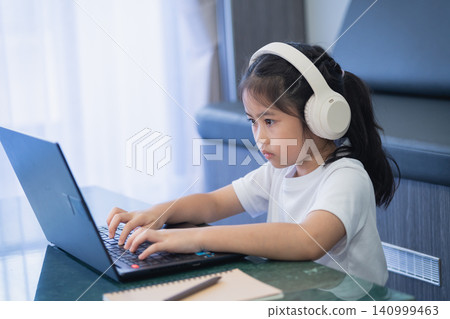 Young girl focused on laptop computer wearing large headphones while sitting at desk indoors in a cozy home workspace or study area with natural light and drapes Young girl focused on laptop computer wearing large headphones while sitting at desk indoors in a cozy home workspace or study area with natural light and drapes 140999463