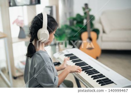 Young girl with braided hair wearing white headphones practicing piano keyboard indoors at home in cozy living room environment Young girl with braided hair wearing white headphones practicing piano keyboard indoors at home in cozy living room environment 140999467
