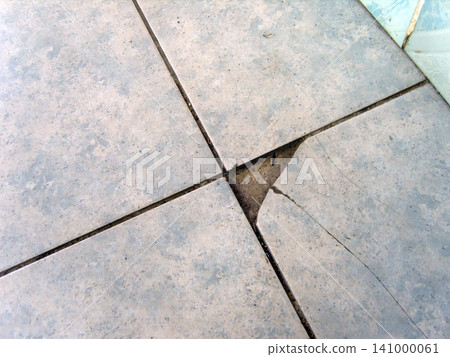 An overhead close-up shot of damaged ceramic floor tiles with broken corner at their intersection. The chipped light gray tile reveals concrete underneath, showing cracked surface needing repair An overhead close-up shot of damaged ceramic floor tiles with broken corner at their intersection. The chipped light gray tile reveals concrete underneath, showing cracked surface needing repair 141000061