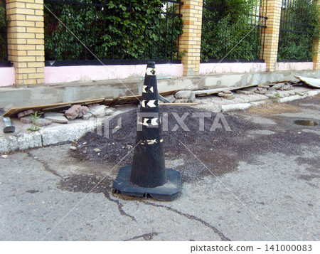 Black traffic cone with white arrows stands on an asphalt street next to a pile of gravel. In the background, a brick fence with a black metal railing and construction debris are visible Black traffic cone with white arrows stands on an asphalt street next to a pile of gravel. In the background, a brick fence with a black metal railing and construction debris are visible 141000083