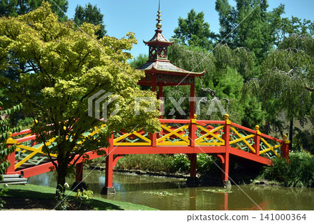 Traditional red Japanese bridge crossing a peaceful pond in a beautiful botanical garden. Scenic landscape with lush trees, calm water and elegant oriental architecture on a sunny day. Traditional red Japanese bridge crossing a peaceful pond in a beautiful botanical garden. Scenic landscape with lush trees, calm water and elegant oriental architecture on a sunny day. 141000364