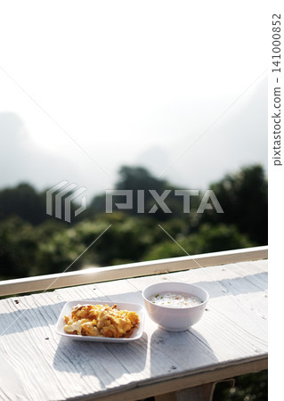 Rice soup and omelet in white plate on white wooden counter bar outdoor is breakfast in resort on the mountain in Thailand Rice soup and omelet in white plate on white wooden counter bar outdoor is breakfast in resort on the mountain in Thailand 141000852