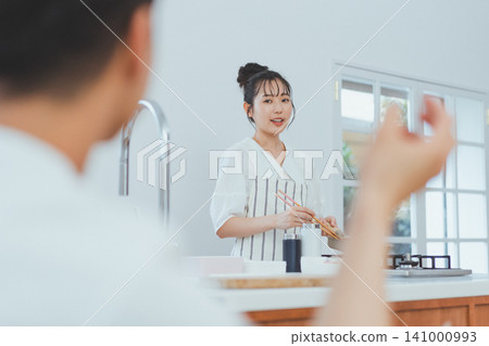 一對情侶在中島廚房做飯的場景:女友面帶微笑,一邊拿著筷子一邊和男友說話。 一對情侶在中島廚房做飯的場景:女友面帶微笑,一邊拿著筷子一邊和男友說話。 141000993