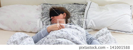 A 50-year-old woman is comfortably sleeping in her bed with a sleep mask on. She is wrapped in a soft blanket, resting peacefully in a serene bedroom setting, banner A 50-year-old woman is comfortably sleeping in her bed with a sleep mask on. She is wrapped in a soft blanket, resting peacefully in a serene bedroom setting, banner 141001588