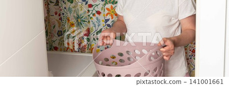 A woman stands in a laundry room holding a basket with clean clothes. She looks focused as she prepares to take out laundry from the washing machine. The room has bright floral wallpaper, banner A woman stands in a laundry room holding a basket with clean clothes. She looks focused as she prepares to take out laundry from the washing machine. The room has bright floral wallpaper, banner 141001661