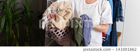 A person stands in a room holding a basket full of dirty clothes. Garments hang in the background. The setting is bright and casual, focusing on the laundry task ahead, banner A person stands in a room holding a basket full of dirty clothes. Garments hang in the background. The setting is bright and casual, focusing on the laundry task ahead, banner 141001662