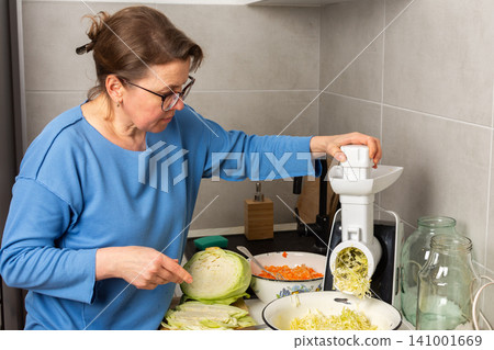 A woman processes cabbage in her kitchen using a food processor. She focuses on shredding the cabbage while other ingredients sit on the counter and in bowls nearby A woman processes cabbage in her kitchen using a food processor. She focuses on shredding the cabbage while other ingredients sit on the counter and in bowls nearby 141001669