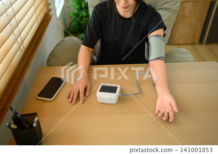 Young Woman Measuring Blood Pressure at Home with Digital Sphygmomanometer and Smartphone on Wooden Table Young Woman Measuring Blood Pressure at Home with Digital Sphygmomanometer and Smartphone on Wooden Table 141001853