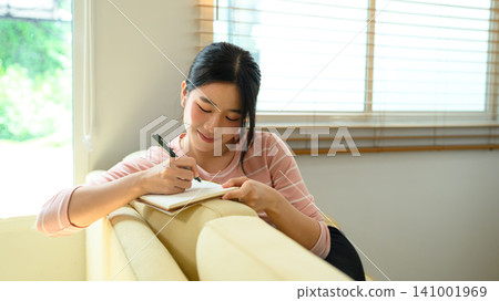 Young Asian Woman Smiling While Writing in Notebook on Sofa at Home 141001969