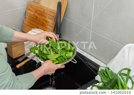 Hands are holding a colander filled with fresh organic local vegetables under running water in a kitchen sink. This is done to clean the produce before cooking 141002010