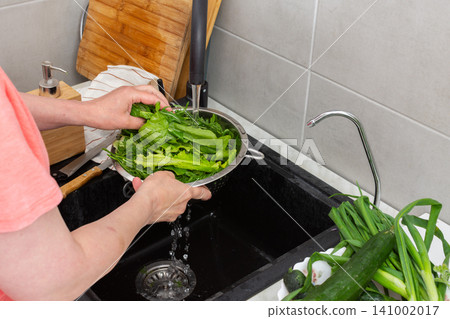 Hands are holding a colander filled with fresh organic local vegetables under running water in a kitchen sink. This is done to clean the produce before cooking Hands are holding a colander filled with fresh organic local vegetables under running water in a kitchen sink. This is done to clean the produce before cooking 141002017