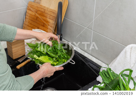 Hands are holding a colander filled with fresh organic local vegetables under running water in a kitchen sink. This is done to clean the produce before cooking Hands are holding a colander filled with fresh organic local vegetables under running water in a kitchen sink. This is done to clean the produce before cooking 141002019