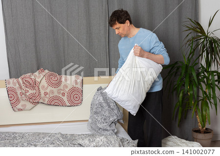 A 53-year-old man is changing the bed linen in his bedroom. He holds a pillow while arranging the sheets and blankets, focusing on the task of keeping his space tidy A 53-year-old man is changing the bed linen in his bedroom. He holds a pillow while arranging the sheets and blankets, focusing on the task of keeping his space tidy 141002077