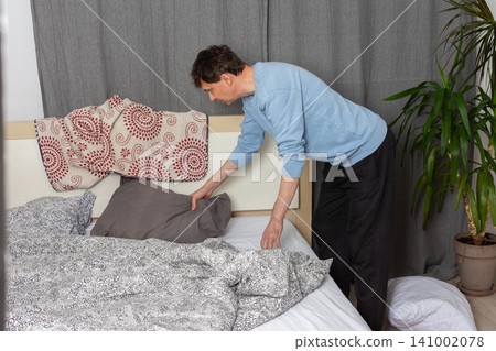 A 53-year-old man replaces the bed linen in his bedroom. He gathers clean sheets and works on making the bed comfortable for sleeping A 53-year-old man replaces the bed linen in his bedroom. He gathers clean sheets and works on making the bed comfortable for sleeping 141002078