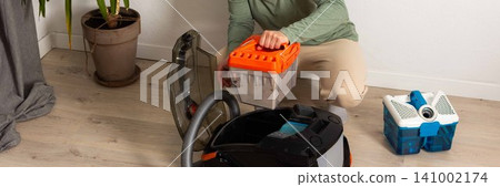 A person is preparing a vacuum cleaner for use by removing the dust container while sitting on the floor. A potted plant is nearby. The room looks bright and tidy, banner A person is preparing a vacuum cleaner for use by removing the dust container while sitting on the floor. A potted plant is nearby. The room looks bright and tidy, banner 141002174