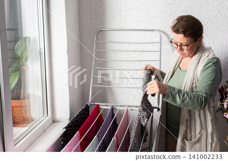 A 50-year-old woman is hanging her laundry on a metal drying rack on her balcony. She looks focused while handling the clothes and has flowers nearby A 50-year-old woman is hanging her laundry on a metal drying rack on her balcony. She looks focused while handling the clothes and has flowers nearby 141002233