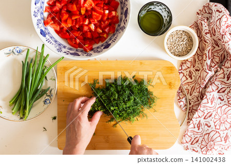 A person is chopping herbs on a wooden board while other fresh vegetables are arranged on a table. Bowls of diced tomatoes and salad dressing are also present 141002438