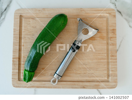 Top view of a fresh green organic zucchini and a metal vegetable peeler placed on a wooden cutting board over a marble surface. Preparing fresh homemade food concept. Cooking healthy ingredient for Top view of a fresh green organic zucchini and a metal vegetable peeler placed on a wooden cutting board over a marble surface. Preparing fresh homemade food concept. Cooking healthy ingredient for 141002507