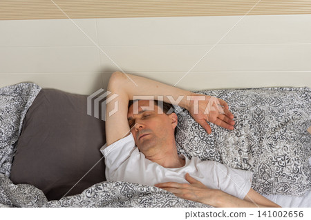 A 53-year-old man is asleep in his bedroom. He is lying on a bed covered with patterned bedding. His arm is stretched above his head. The room is quiet and inviting A 53-year-old man is asleep in his bedroom. He is lying on a bed covered with patterned bedding. His arm is stretched above his head. The room is quiet and inviting 141002656