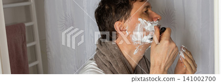 A 53 year old man shaves in the bathroom during his morning routine. He uses a razor and shaving cream while wearing a towel around his neck. Water is visible in the sink, banner A 53 year old man shaves in the bathroom during his morning routine. He uses a razor and shaving cream while wearing a towel around his neck. Water is visible in the sink, banner 141002700