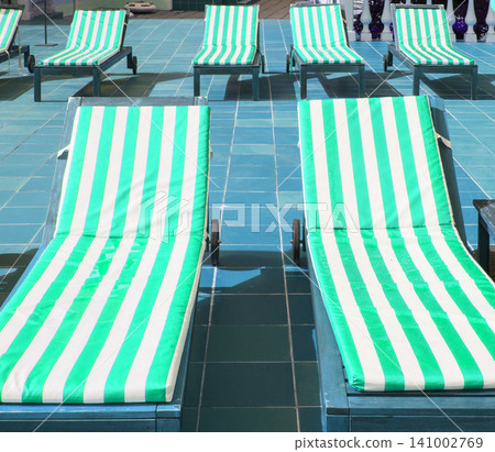 Empty wooden deck chairs with mattresses in green and white stripes stand in a row. Empty wooden deck chairs with mattresses in green and white stripes stand in a row. 141002769