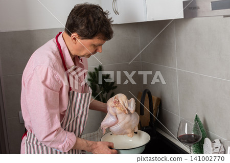 In a cozy kitchen, a 53-year-old man washes chicken in the sink while getting ready for a pre-Christmas dinner. He has potatoes and fruits on the table beside him In a cozy kitchen, a 53-year-old man washes chicken in the sink while getting ready for a pre-Christmas dinner. He has potatoes and fruits on the table beside him 141002770