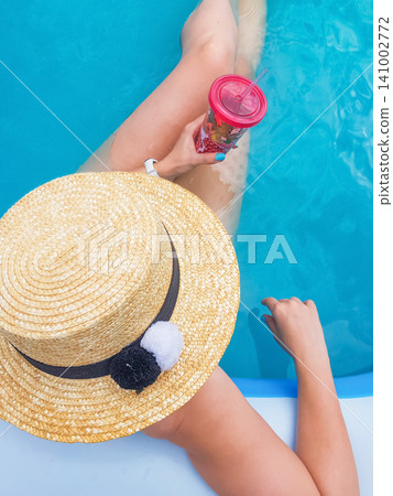 A stylish lady in a straw hat is relaxing in the pool water and holding a cocktail glass top view 141002772