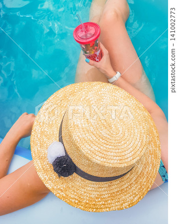 A stylish lady in a straw hat is relaxing in the pool water and holding a cocktail glass A stylish lady in a straw hat is relaxing in the pool water and holding a cocktail glass 141002773
