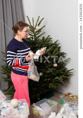 A person adds white ornaments to a Christmas tree. The tree is standing in a living room with gray curtains. The activity happens during the holiday season at home A person adds white ornaments to a Christmas tree. The tree is standing in a living room with gray curtains. The activity happens during the holiday season at home 141002850