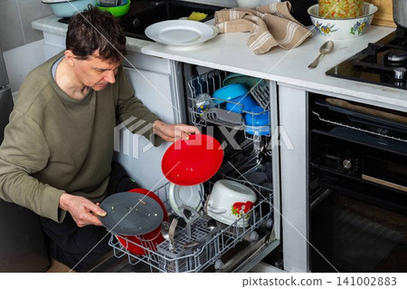 A man is putting dirty dishes into the dishwasher in his kitchen. He focuses on the task while several items rest in the appliance, ready for cleaning A man is putting dirty dishes into the dishwasher in his kitchen. He focuses on the task while several items rest in the appliance, ready for cleaning 141002883
