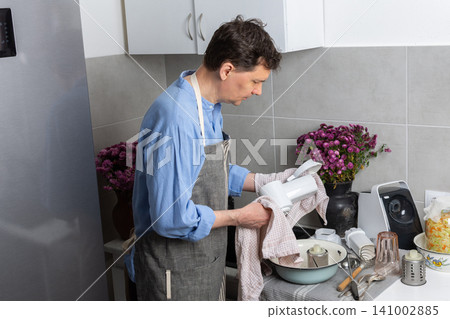 A man stands in the kitchen wiping clean dishes with a cloth. He wears an apron and focuses on his task. The kitchen has a sink and some dishes on the counter A man stands in the kitchen wiping clean dishes with a cloth. He wears an apron and focuses on his task. The kitchen has a sink and some dishes on the counter 141002885