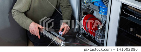 In a kitchen, a man is putting a dishwashing detergent tablet into the dishwasher. He is getting ready to clean the dirty dishes inside the machine, banner In a kitchen, a man is putting a dishwashing detergent tablet into the dishwasher. He is getting ready to clean the dirty dishes inside the machine, banner 141002886