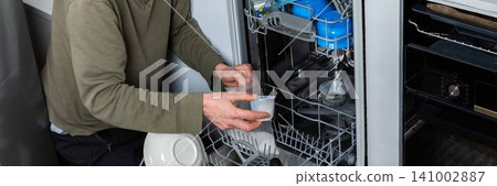 A man is at work in the kitchen, pouring salt into a dishwasher as he prepares to wash dirty dishes. He is focused on the task at hand while organizing the appliances, banner A man is at work in the kitchen, pouring salt into a dishwasher as he prepares to wash dirty dishes. He is focused on the task at hand while organizing the appliances, banner 141002887