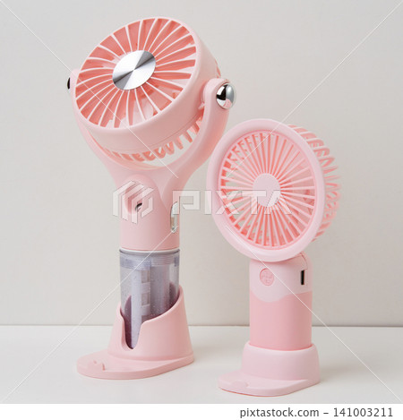 Two pink portable mini fans standing on a white surface against a neutral light background, summer cooling gadget concept Two pink portable mini fans standing on a white surface against a neutral light background, summer cooling gadget concept 141003211