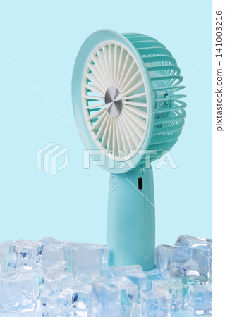 Blue portable mini fan standing among ice cubes on light blue background, extreme summer cooling concept Blue portable mini fan standing among ice cubes on light blue background, extreme summer cooling concept 141003216