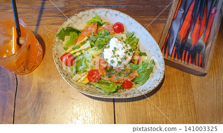 Appetizing salad with salmon, cherry tomatoes, herbs and stracciatella cheese on a wooden table next to a refreshing 141003325