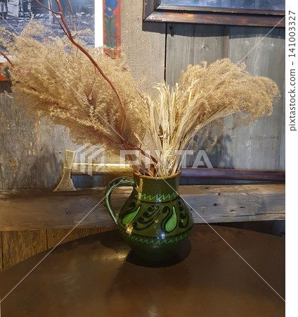 Ethnic green ceramic jug with dried flowers and wheat ears on a round table in an authentic interior with wooden walls Ethnic green ceramic jug with dried flowers and wheat ears on a round table in an authentic interior with wooden walls 141003327