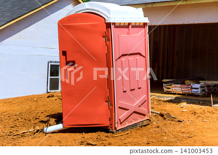 Portable toilet stands alone on dirt ground next to building site with construction materials. Portable toilet stands alone on dirt ground next to building site with construction materials. 141003453