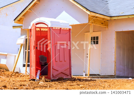 Portable toilet sits outside house under construction with dirt around area during work hours. Portable toilet sits outside house under construction with dirt around area during work hours. 141003471