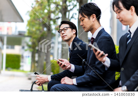 A man in a suit sits on a bus stop bench, waiting for the bus. A man in a suit sits on a bus stop bench, waiting for the bus. 141003830