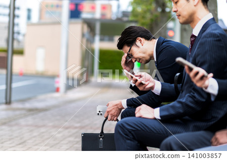 A man in a suit sits dejectedly on a bus stop bench. 141003857