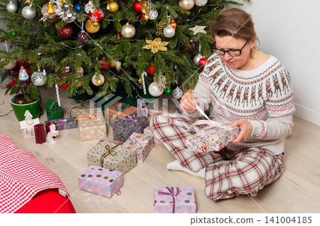 A woman aged 52 sits on the floor next to a decorated Christmas tree. She opens gifts on Christmas Day while enjoying the holiday with family members in the background A woman aged 52 sits on the floor next to a decorated Christmas tree. She opens gifts on Christmas Day while enjoying the holiday with family members in the background 141004185