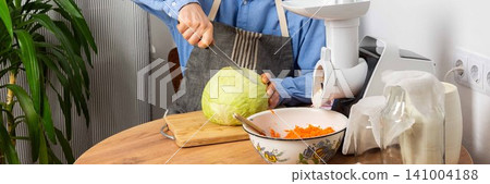 A 53-year-old man cuts cabbage with a knife while preparing to ferment cabbage and carrots. He stands in his kitchen near a vegetable grinder and a bowl of grated carrots, banner 141004188