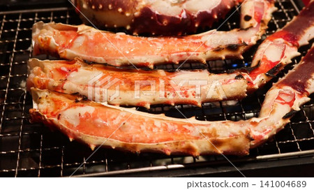 close up of four Grilled king crab legs sizzling on a metal wire rack over an open flame. These split crab legs showcase tender, juicy white and pink meat with beautiful char marks from grilling. close up of four Grilled king crab legs sizzling on a metal wire rack over an open flame. These split crab legs showcase tender, juicy white and pink meat with beautiful char marks from grilling. 141004689