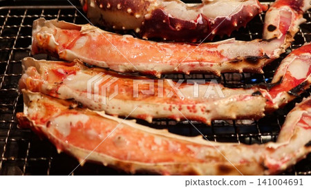 close up of  four Grilled king crab legs sizzling on a metal wire rack over an open flame. These split crab legs showcase tender, juicy white and pink meat with beautiful char marks from grilling. 141004691