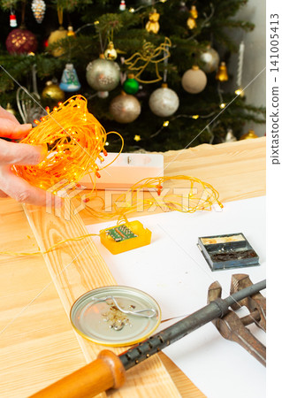 A man fixes a Christmas tree garland and lights on a table at home. He is checking the connection and making adjustments to ensure everything works for the holiday season A man fixes a Christmas tree garland and lights on a table at home. He is checking the connection and making adjustments to ensure everything works for the holiday season 141005413
