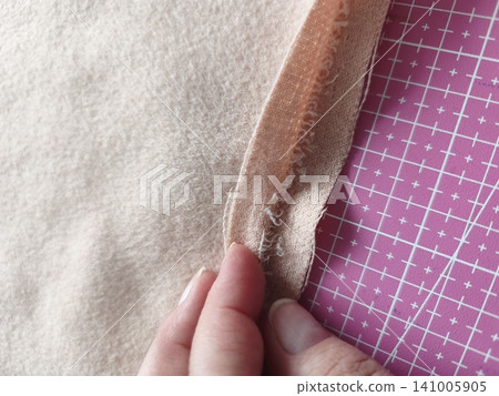 A top-down view of a seamstress or tailor hand-sewing and finishing the edge of a beige double-faced coat fabric. A top-down view of a seamstress or tailor hand-sewing and finishing the edge of a beige double-faced coat fabric. 141005905