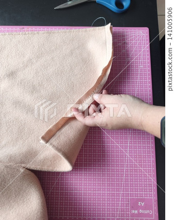 Tailor's hands in the foreground hand stitching the edge of a double-faced wool coat. Tailor's hands in the foreground hand stitching the edge of a double-faced wool coat. 141005906