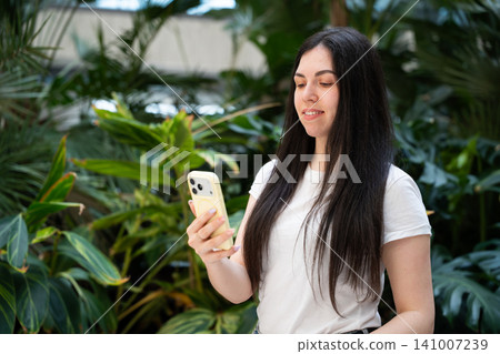 Young woman using smartphone for digital communication in green office environment. Mobile app technology remote work and sustainable workplace 141007239