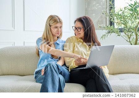 Happy mother and teenage daughter pointing at laptop screen together on sofa Happy mother and teenage daughter pointing at laptop screen together on sofa 141007362